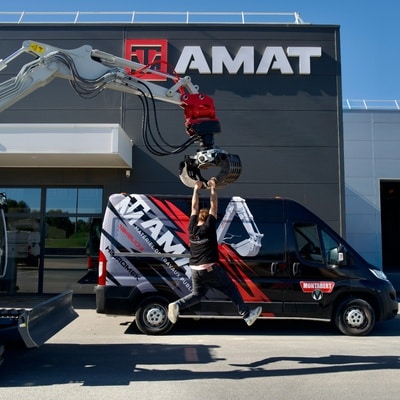 Homme suspendu au grappin d'une pelle devant le bâtiment et un fourgon AMAT Matériel de Travaux Publics.