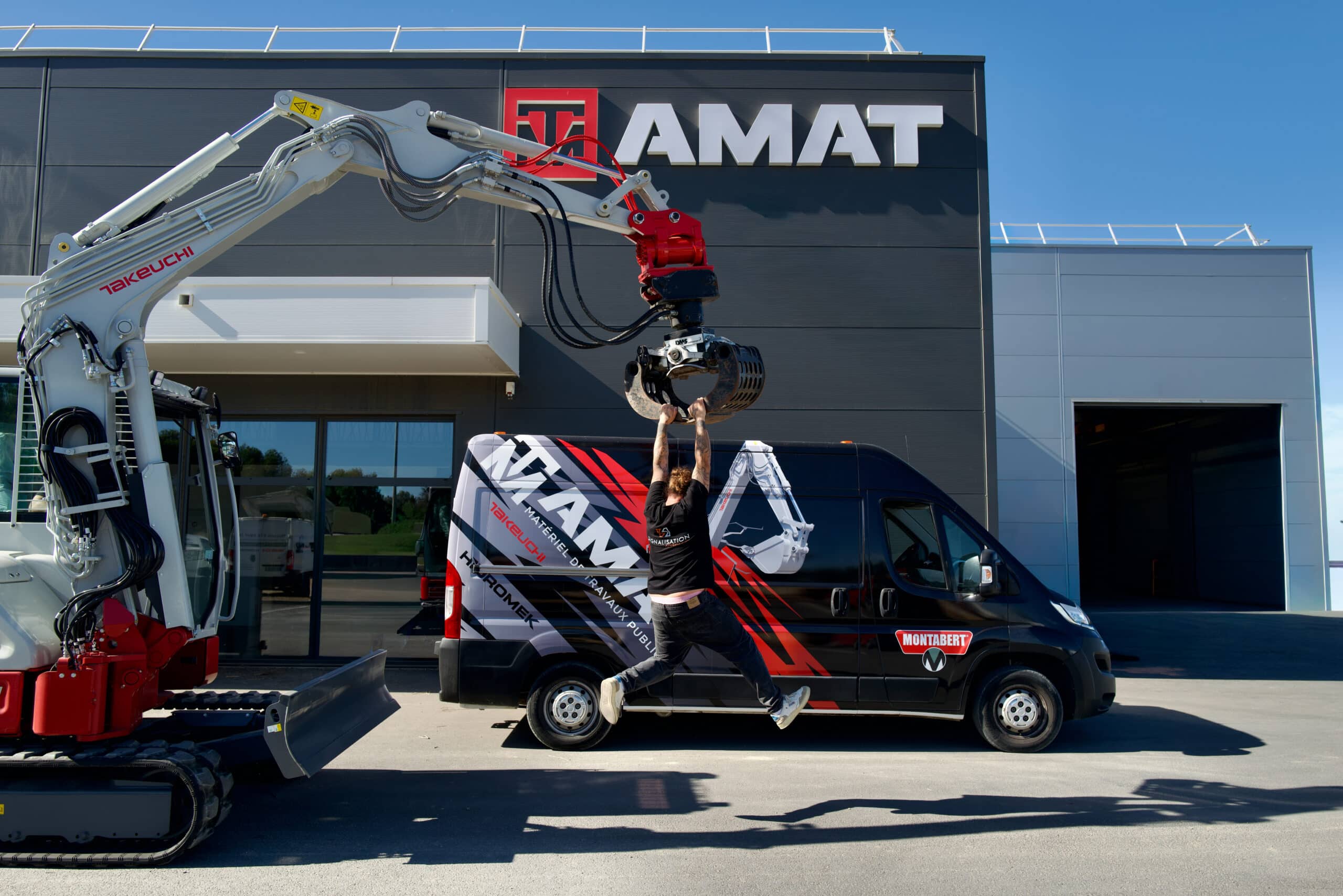 AMAT : Matériel de Travaux Publics et Pelleteuse Takeuchi Homme suspendu au grappin d'une pelle Takeuchi devant l'enseigne AMAT et un van de matériel de travaux publics.
