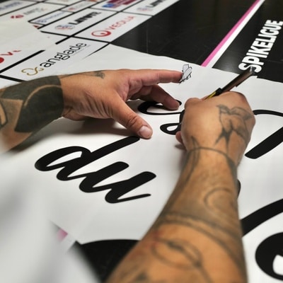 Mains tatouées réalisant une calligraphie sur une affiche Spikeligue devant divers logos de partenaires.