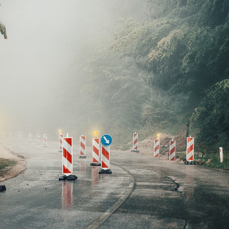 Chantier brumeux : signalisation et barrières sur route mouillée. Route mouillée et brumeuse. Barrières de chantier rayées rouges et blanches avec lumières clignotantes et flèche bleue de direction.