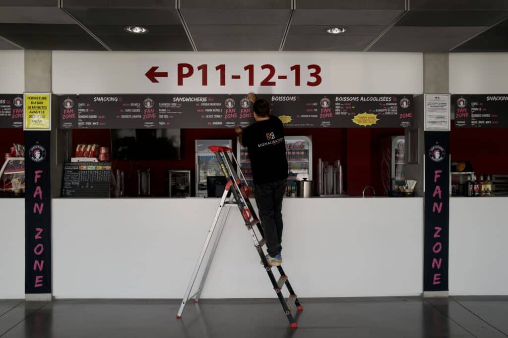 Ouvrier sur escabeau installant un panneau de menu pour la Fan Zone des Centurions de Narbonne, signalétique P11-12-13.