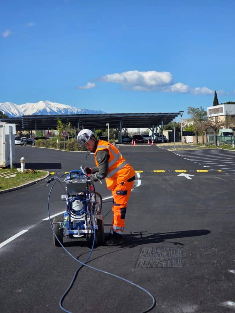Ouvrier utilisant une machine de marquage routier sur un parking, avec des montagnes enneigées en arrière-plan.