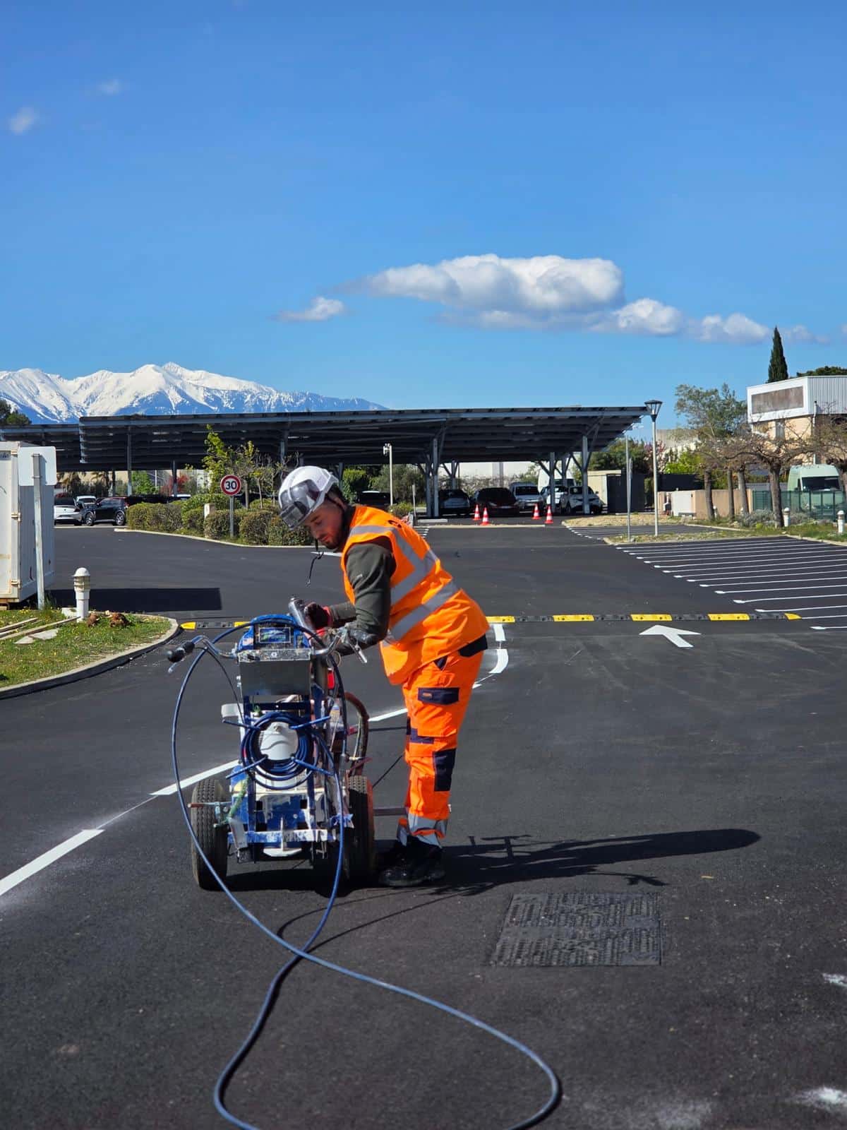 Marquage au sol de parking par un professionnel du traçage Ouvrier utilisant une machine de marquage routier sur un parking, avec des montagnes enneigées en arrière-plan.