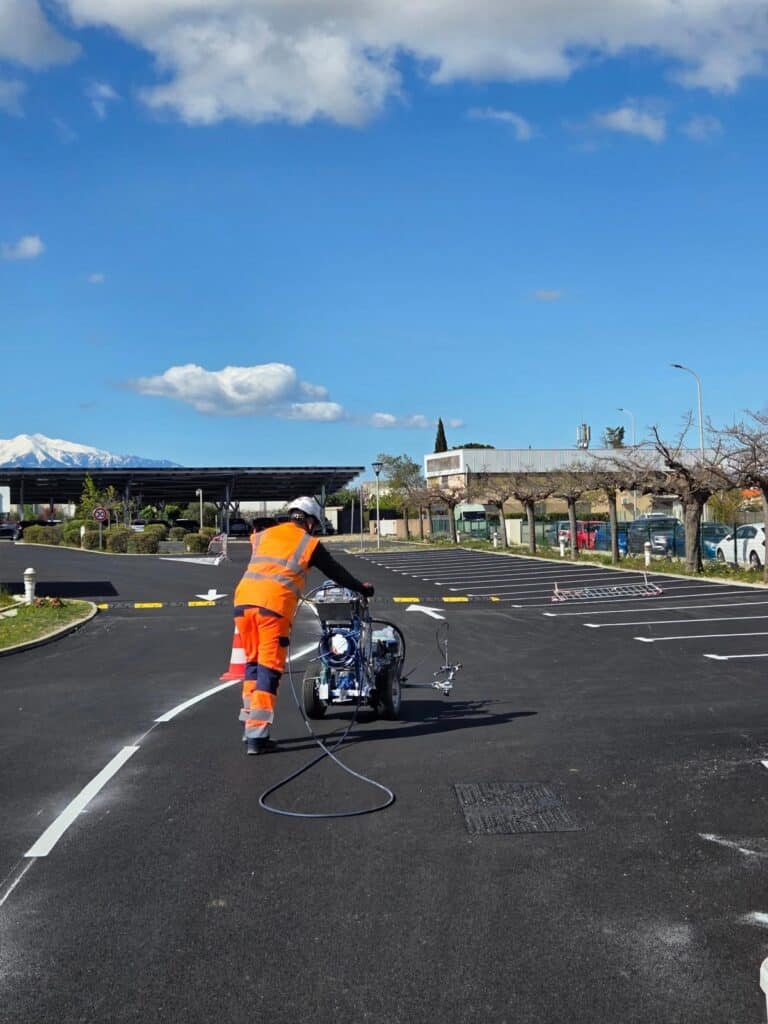 Ouvrier en tenue orange traçant des lignes blanches dans un parking avec une machine de marquage au sol.