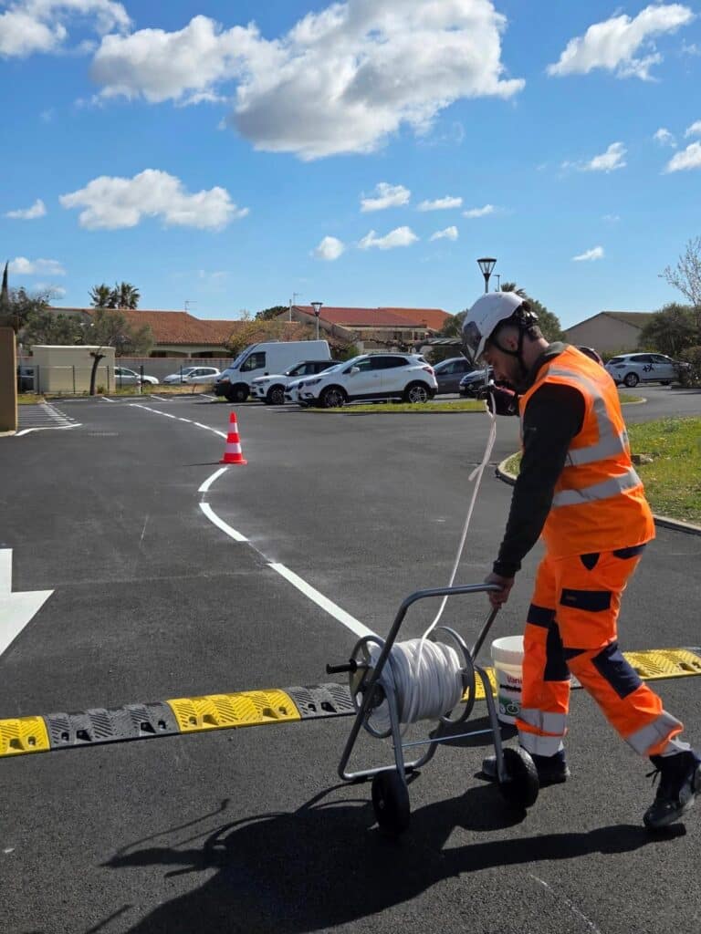 Agent de signalisation traçant une ligne blanche courbe dans un parking avec un applicateur manuel sur bitume.