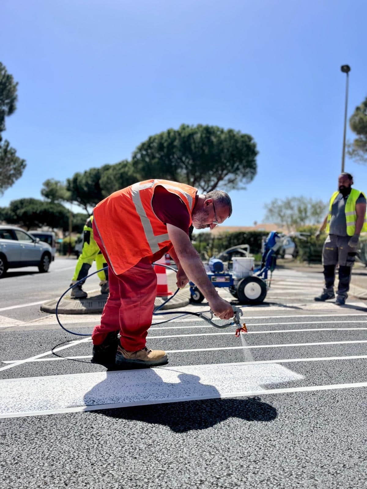 Marquage routier : Ouvrier peignant un passage piéton Ouvrier en gilet orange peignant un passage piéton avec un pistolet pulvérisateur sur une route ensoleillée.