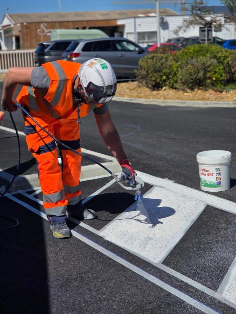 Ouvrier en tenue haute visibilité peignant un marquage au sol Varilux Color SRT sur bitume avec un pistolet.