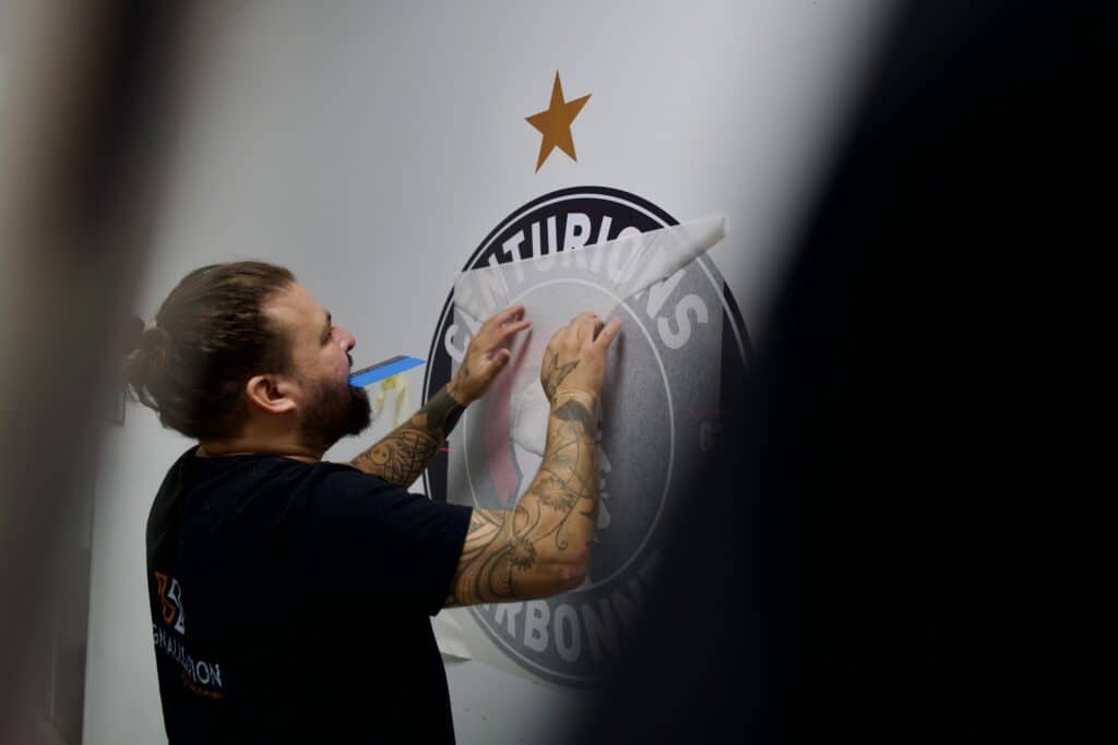 Pose de Sticker Mural Centurions Narbonne - Signalétique Artisan tatoué posant un sticker mural Centurions Narbonne avec précision sur un mur blanc.